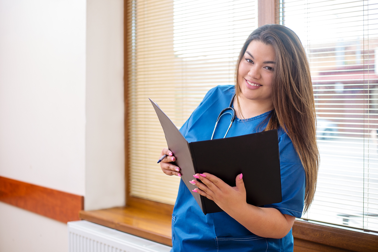 Smiling confident female doctor standing in a hospital hallway next to a window, wearing a blue uniform and stethoscope, holding a folder